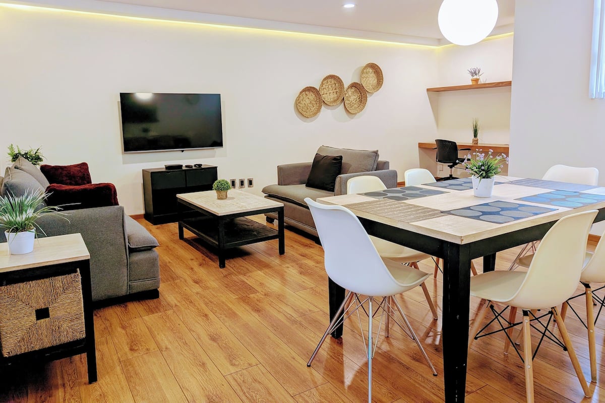 Una sala moderna con un televisor de pantalla plana, un sofá, una mesa de centro y una zona de comedor con una mesa y sillas.