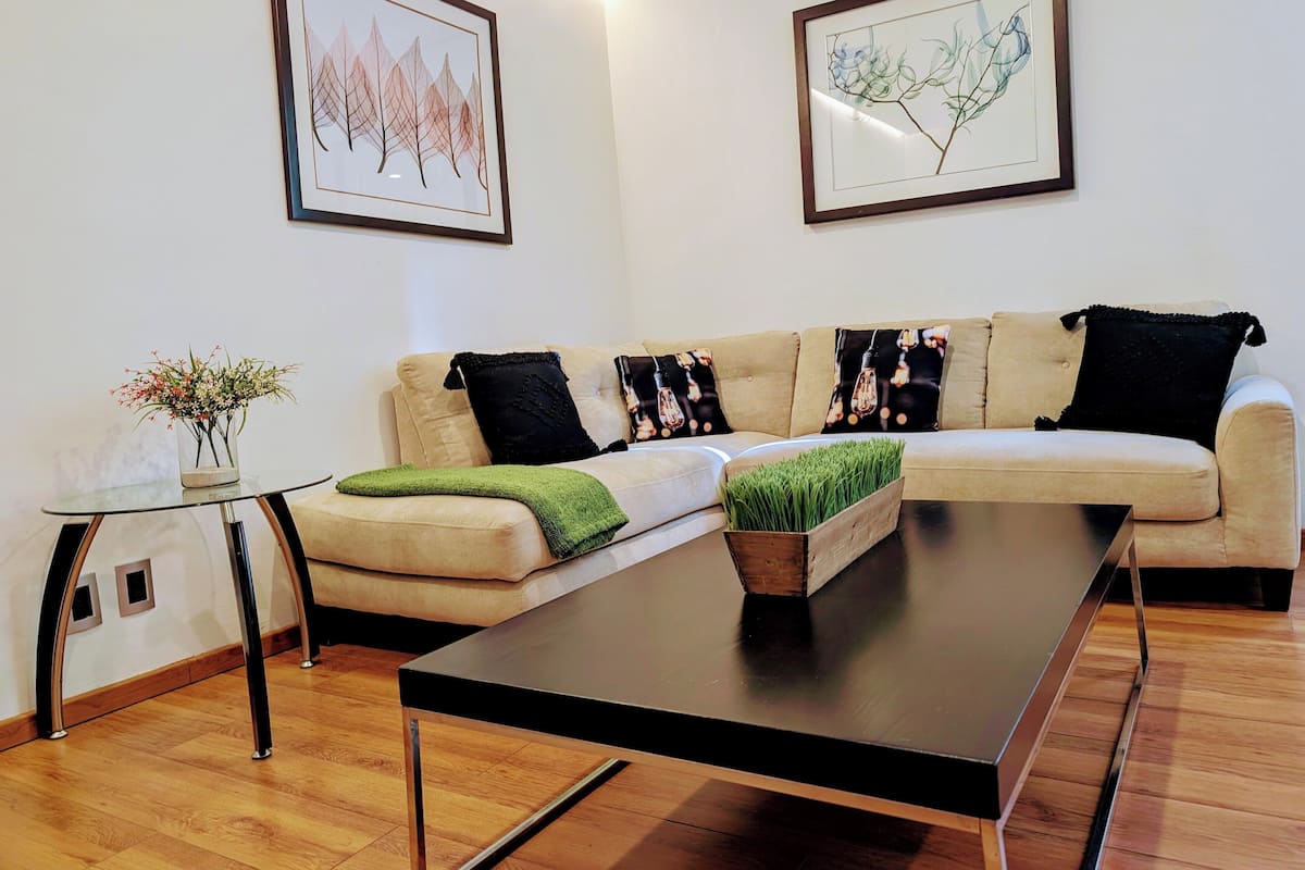 Una sala de estar moderna con un sofá color beige, cojines estampados en blanco y negro, una mesa de centro de vidrio y dos obras de arte enmarcadas en la pared.