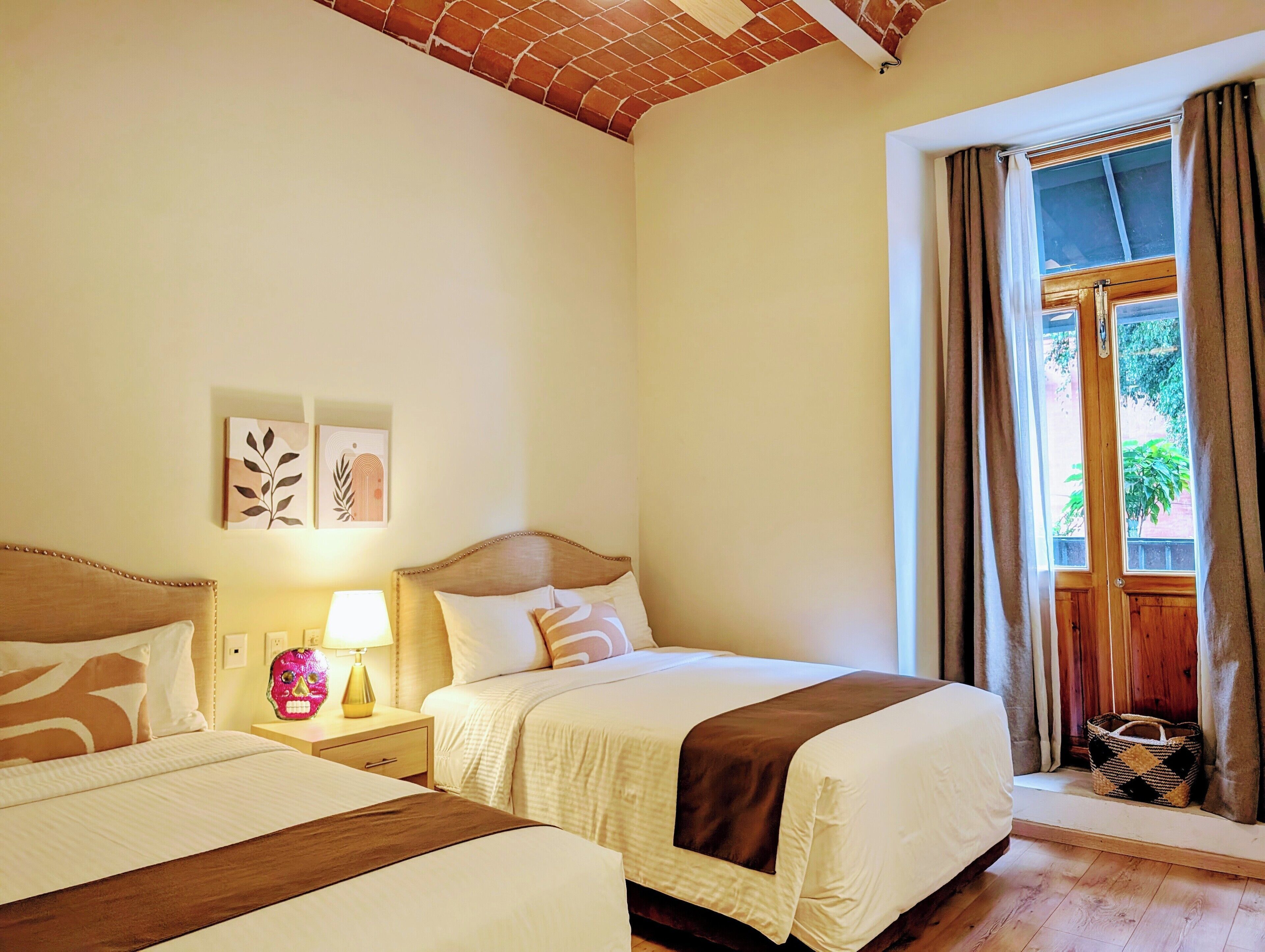 A hotel room with two beds, a wooden headboard, a nightstand with a lamp, artwork on the wall, and a window with curtains.