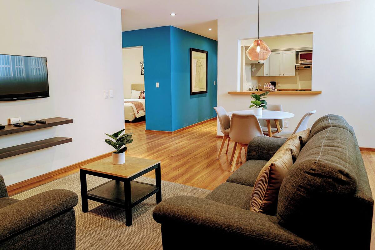 Una sala de estar moderna con un sofá, una mesa de centro y un televisor. Hay una zona de comedor con una mesa y sillas, y una cocina al fondo.