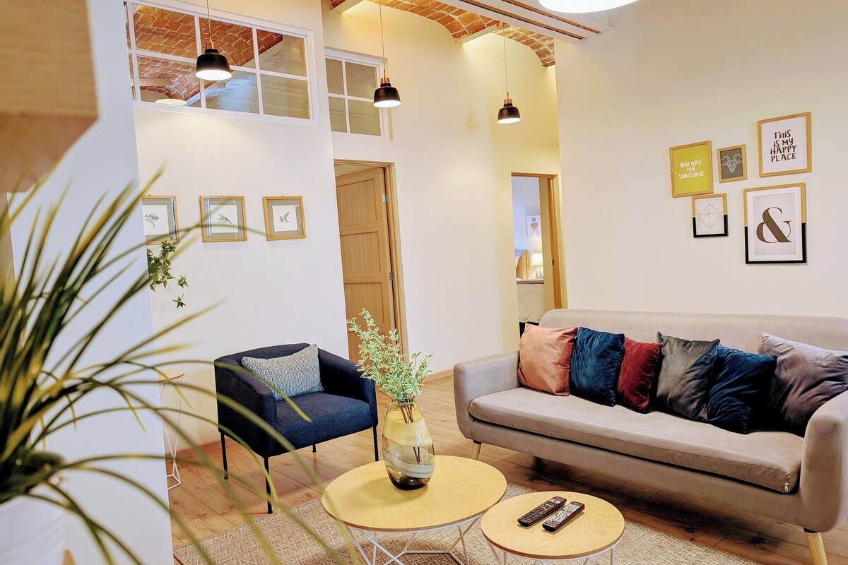 Una sala de estar moderna con un sofá, sillones y una mesita de centro. Hay cuadros enmarcados en la pared y una planta en el suelo.