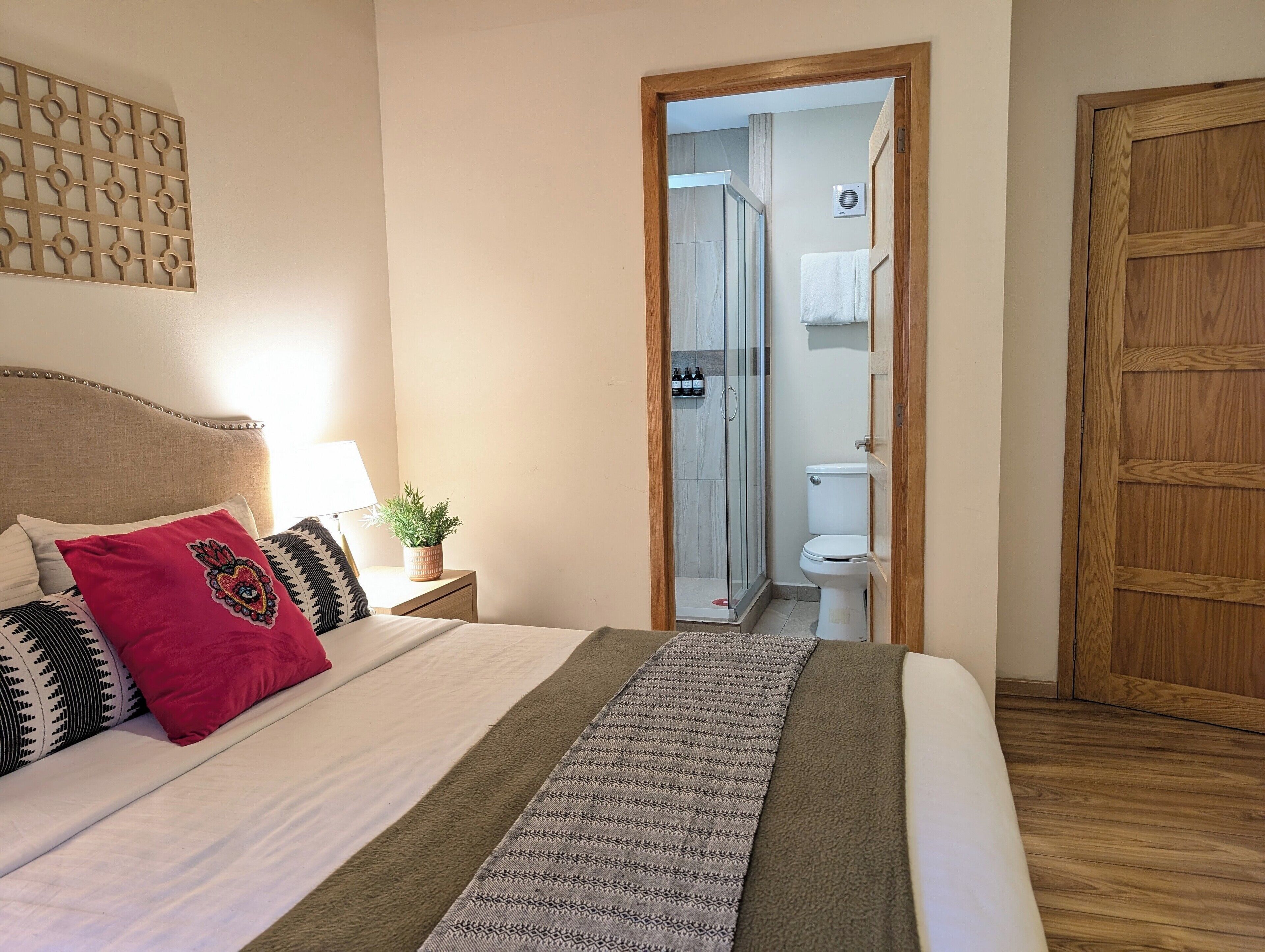 A neatly made bed with a patterned blanket, a bedside table with a lamp and a plant, and a wooden door.