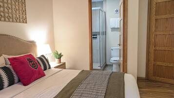 A neatly made bed with a patterned blanket, a bedside table with a lamp and a plant, and a wooden door.