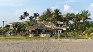 A beachside resort with a thatched-roof hut, palm trees, and a building with a red roof in the background. - Hotel Villa Murano (Tonalá)
