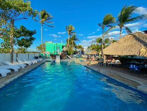 A swimming pool with lounge chairs, palm trees, and a thatched-roof structure.