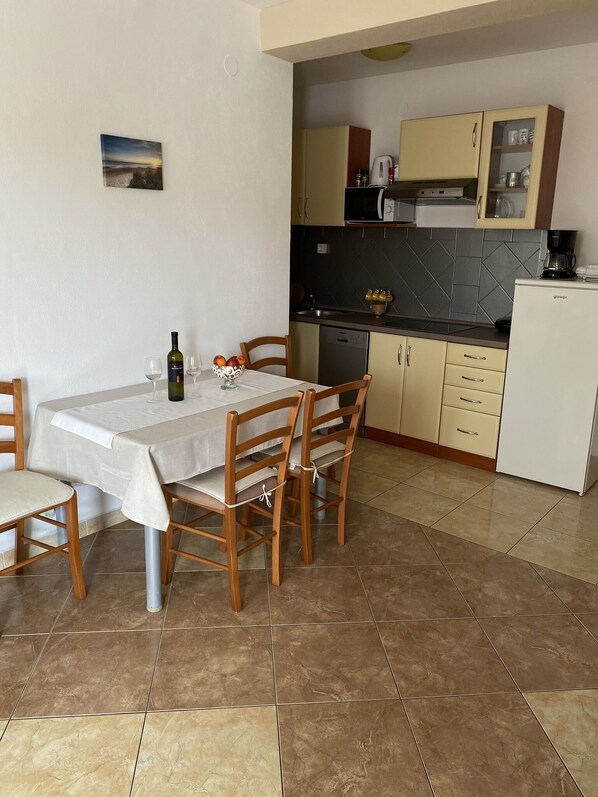 Un comedor con una mesa para dos, sillas de madera, un pequeño cuadro en la pared y una cocina al fondo.