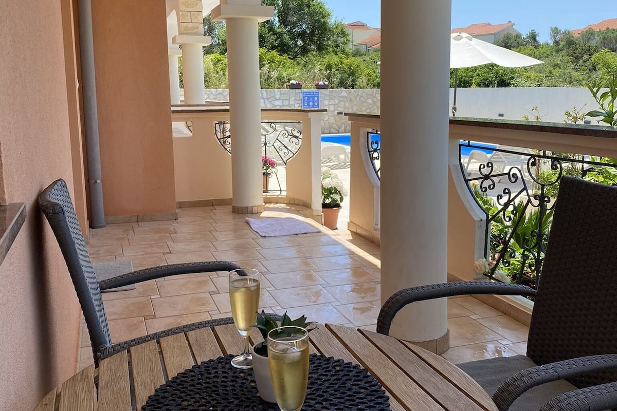 Een houten tafel met twee champagneglazen op een balkon met witte zuilen en een uitzicht op groen en huizen op de achtergrond.