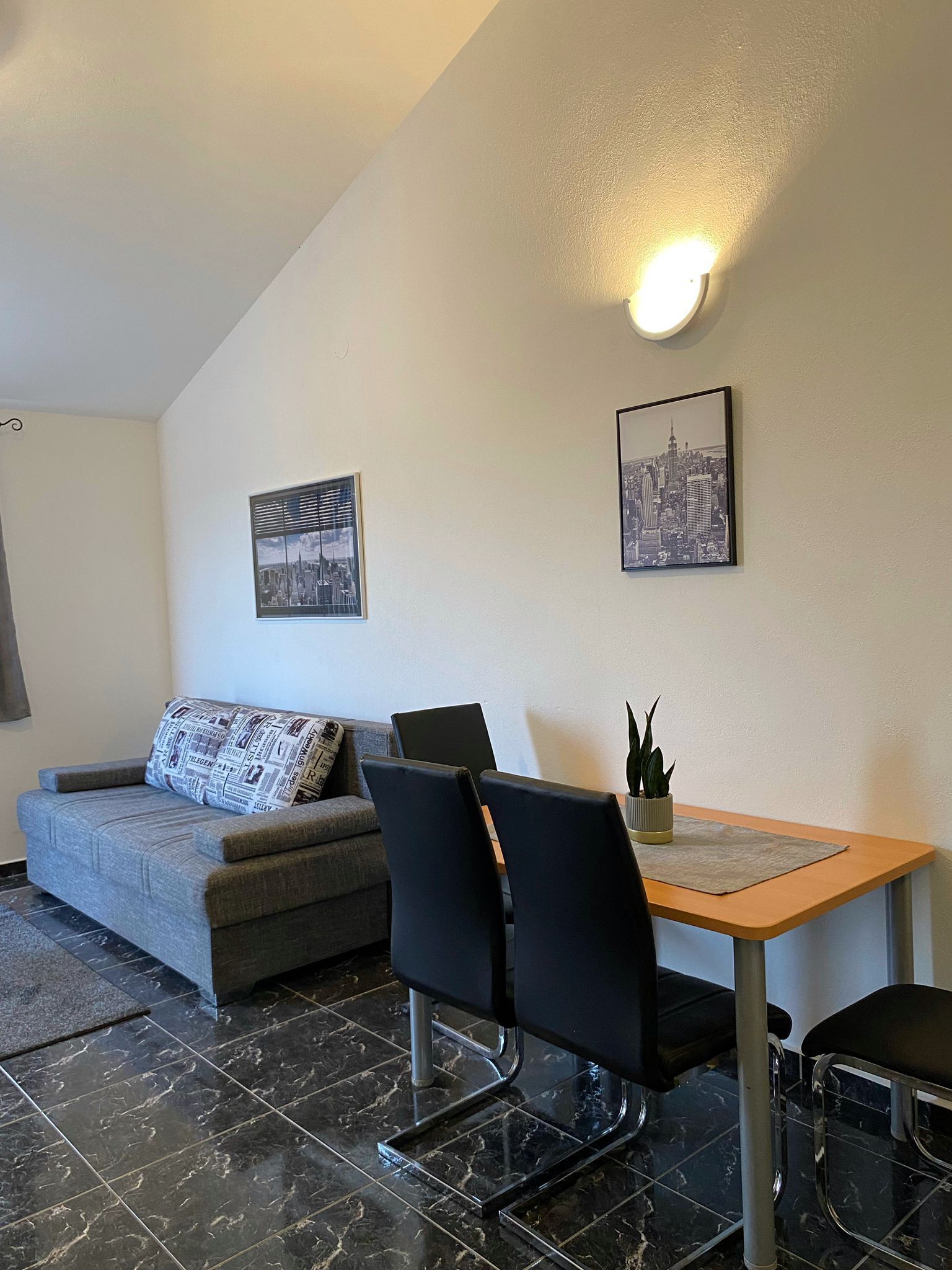 A modern living room with a grey sofa, black chairs, a wooden dining table, and two cityscape wall art pieces.
