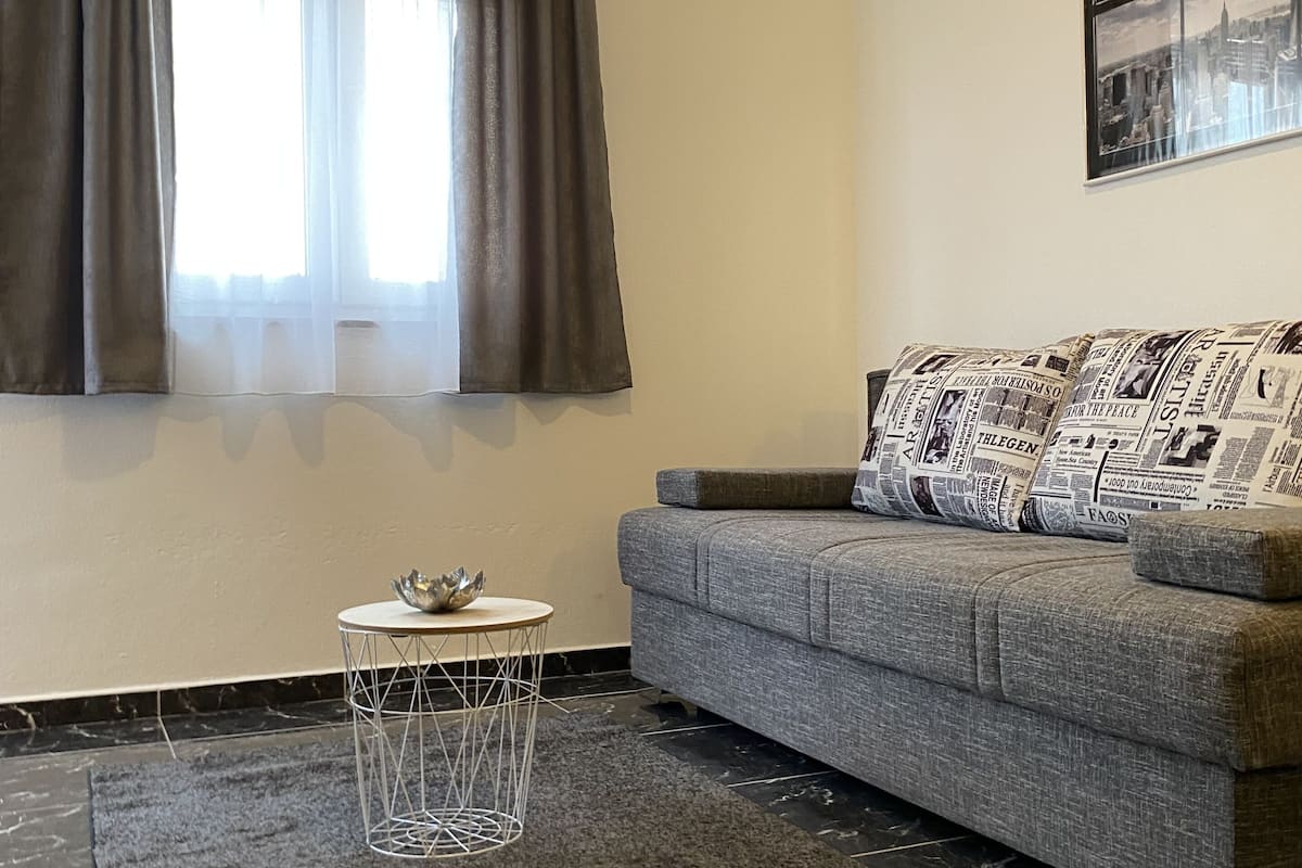 Een woonkamer met een grijze bank, een klein rond tafeltje met een metalen poot, een donkergrijze vloerkleed en een raam met gordijnen.