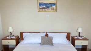 Een net opgemaakt bed met witte lakens, twee nachtkastjes met lampen, een schilderij van een strand en uitzicht op zee.