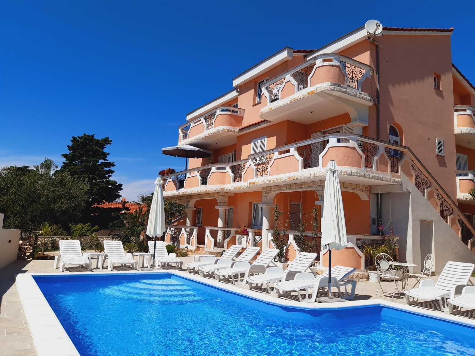 Un hotel de varios pisos con piscina, sillas de descanso blancas y sombrillas para el exterior.