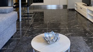 Een moderne woonkamer met een grijze bank, een bijzettafeltje met een decoratieve schaal, een eethoek met een tafel en stoelen, en een keuken met witte kastjes en een betegelde achterwand.