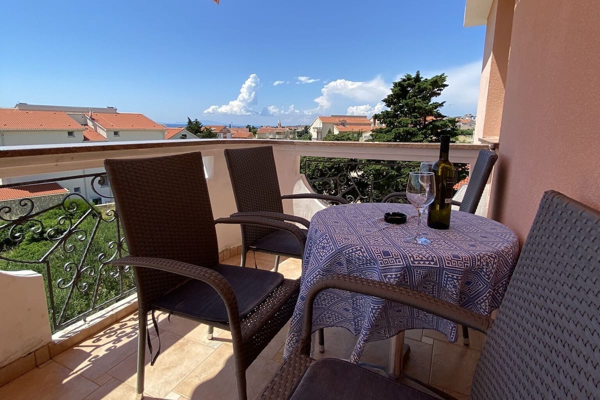 Een balkon met rieten stoelen en een tafel, een gestreepte luifel en uitzicht op gebouwen en bomen.