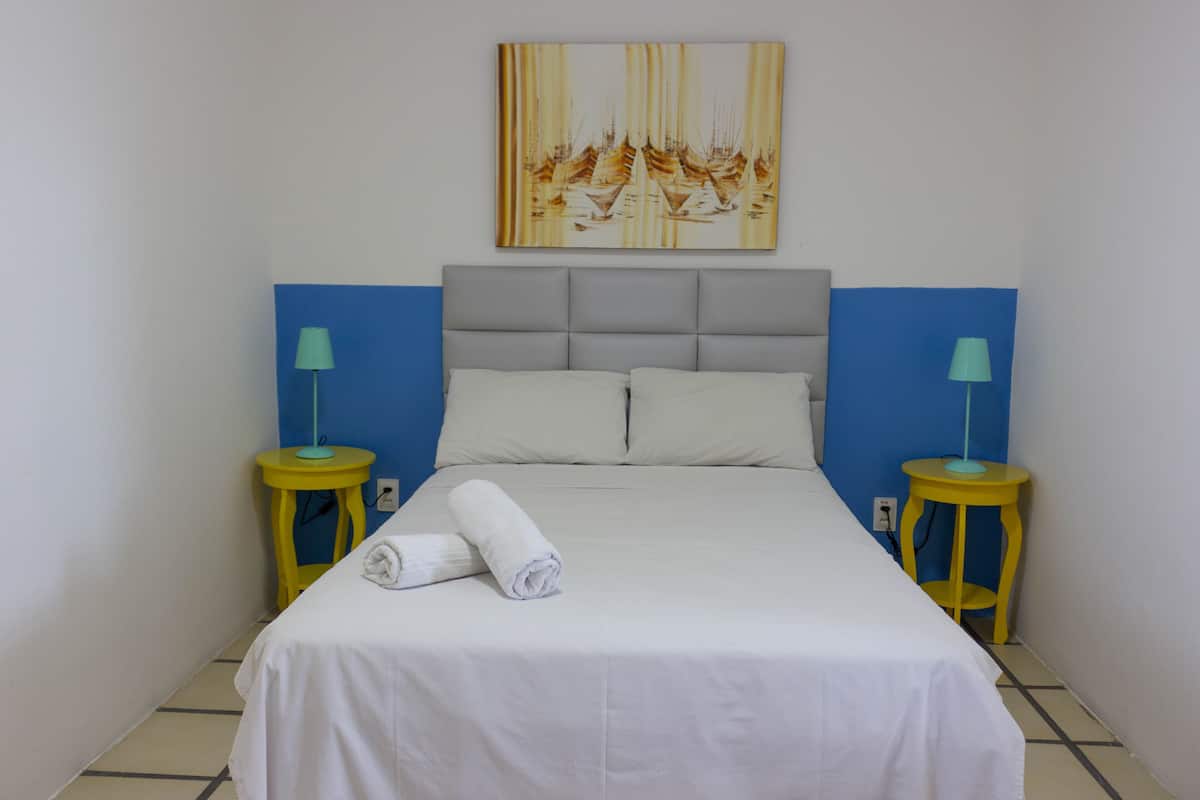 Un lit bien fait, avec du linge de lit blanc et deux serviettes enroulées, flanqué de deux tables de chevet jaunes avec des lampes, dans une pièce aux murs bleus et ornée d’un grand tableau au-dessus du lit.