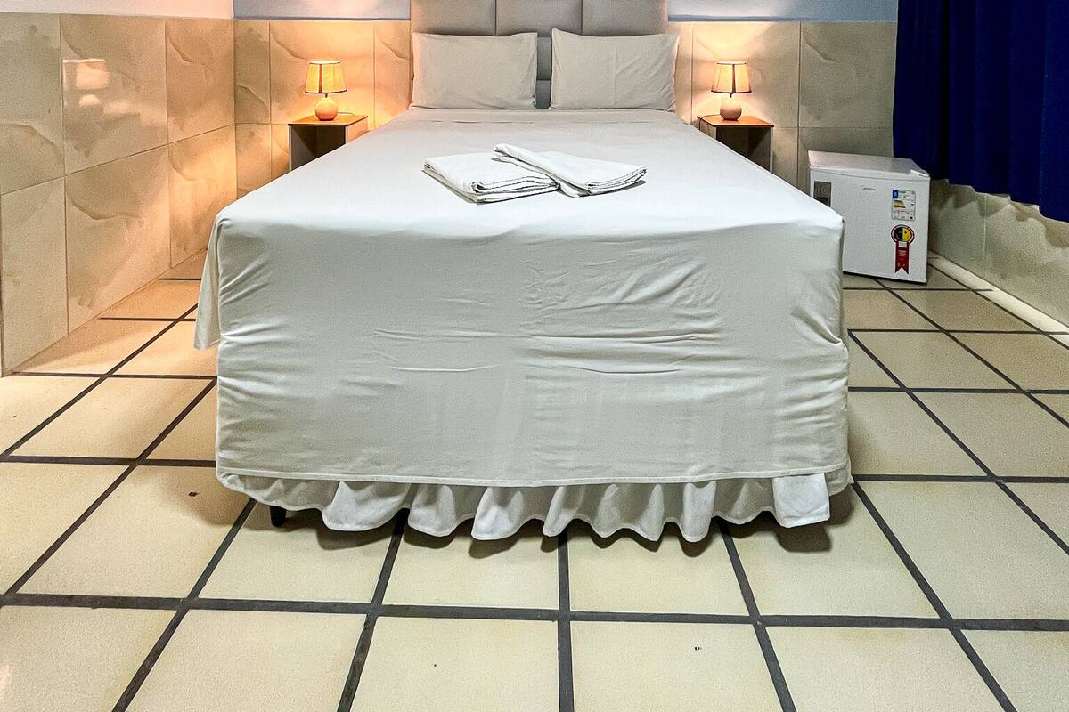 Un lit bien fait, recouvert d’une housse de couette blanche et agrémenté d’oreillers, un climatiseur fixé au mur et une table de chevet avec une lampe.