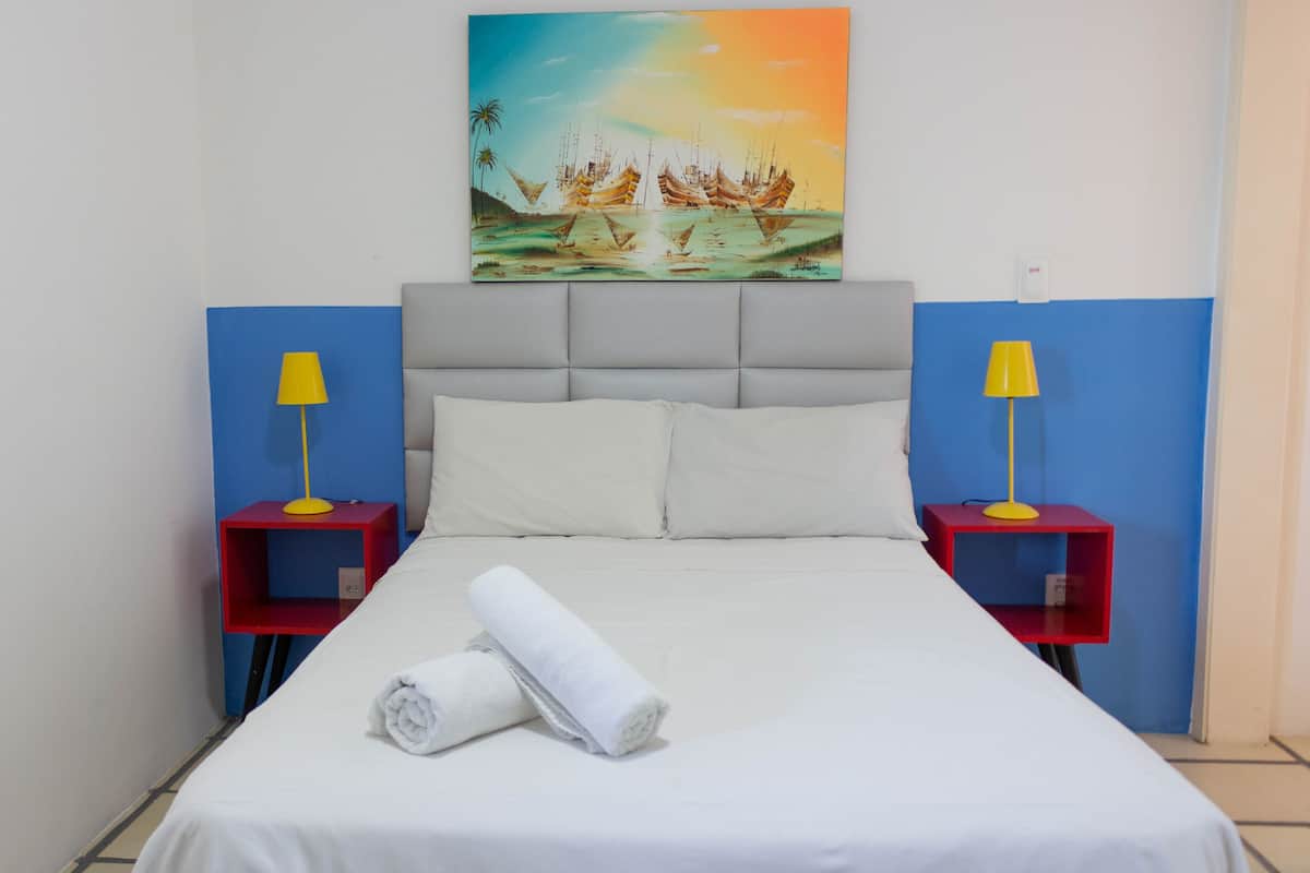 Un lit bien fait, avec du linge de lit blanc et une serviette enroulée, flanqué de deux tables de chevet rouges surmontées de lampes jaunes, dans une pièce dont un mur est peint en bleu et où un tableau représentant des bateaux est accroché au-dessus du lit.