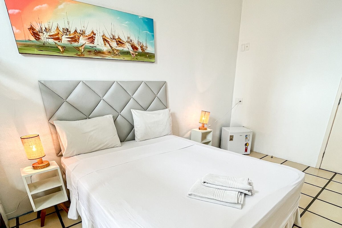 Une chambre d’hôtel bien rangée, avec un grand lit, une table de chevet avec une lampe, un petit radiateur et un tableau accroché au mur.
