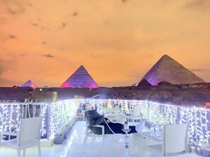 Un área de comedor al aire libre con mesas y sillas, iluminada con luces colgantes, y pirámides al fondo.