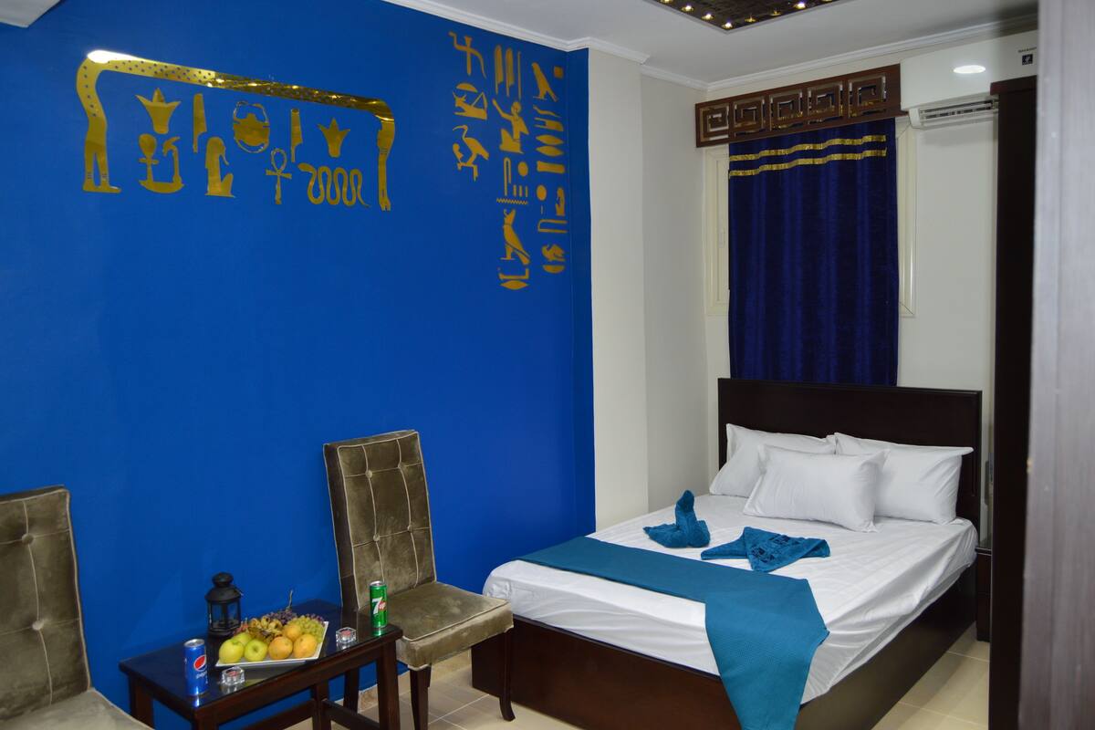 A hotel room with a blue wall, a bed with white linens, a blue throw blanket, a chair, a small table with fruit and a can of soda, and decorative wall art.