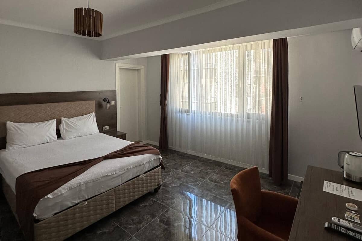 Une chambre d’hôtel avec un lit, un bureau, une chaise, une fenêtre avec des rideaux et un luminaire au plafond.