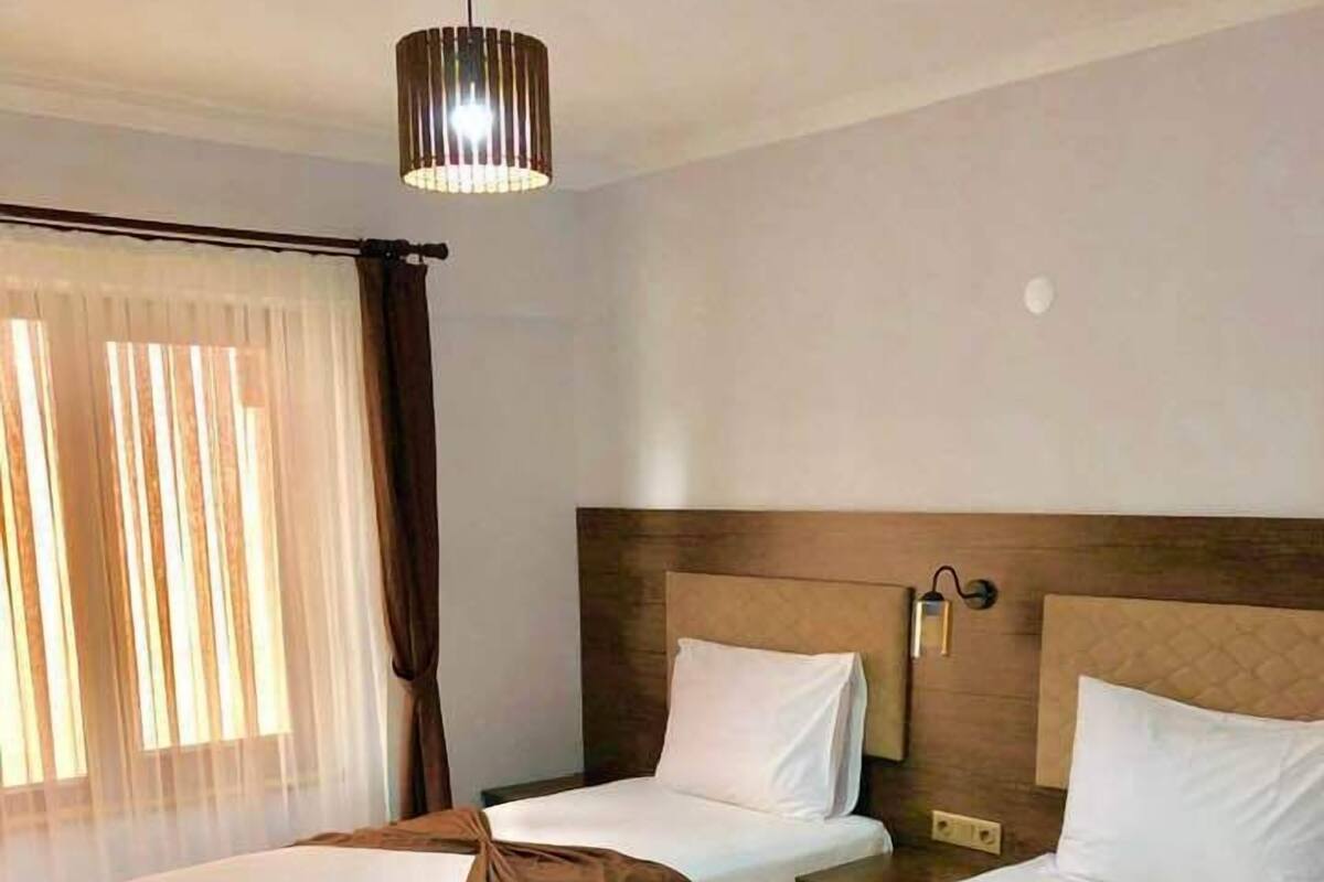Une chambre d’hôtel avec trois lits simples, une tête de lit en bois, un luminaire au plafond et une fenêtre avec des rideaux légers.