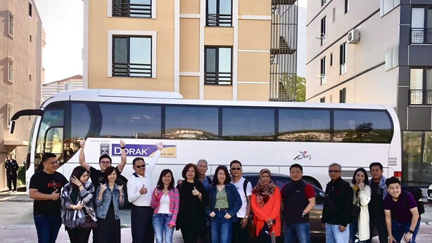 Eine Gruppe von Menschen steht vor einem weißen Bus mit einem DORAK-Schild, der an einer Straße mit modernen Gebäuden im Hintergrund geparkt ist.
