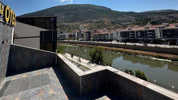 Een hotel met een modern design, met een opvallend ‘HOTEL’ bord, een terras met stenen vloeren en uitzicht op een rivier en omliggende gebouwen.