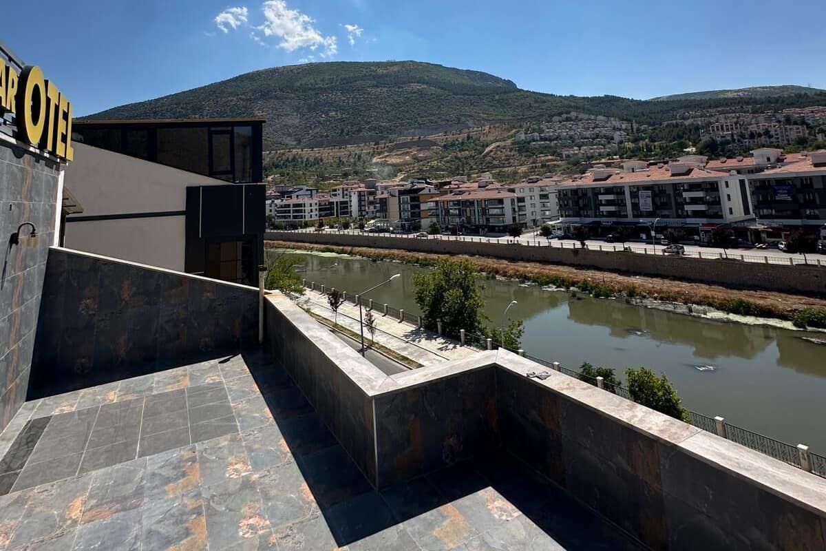 Un hôtel au design moderne, avec une enseigne « HÔTEL » bien visible, une terrasse dotée d’un sol en pierre et offrant une vue sur une rivière et les bâtiments environnants.
