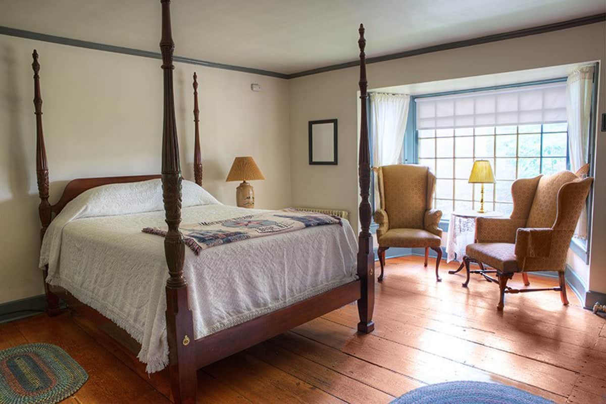 Una cama con dosel, un edredón, dos sillones, un ventanal con cortinas y una lámpara sobre un piso de madera.