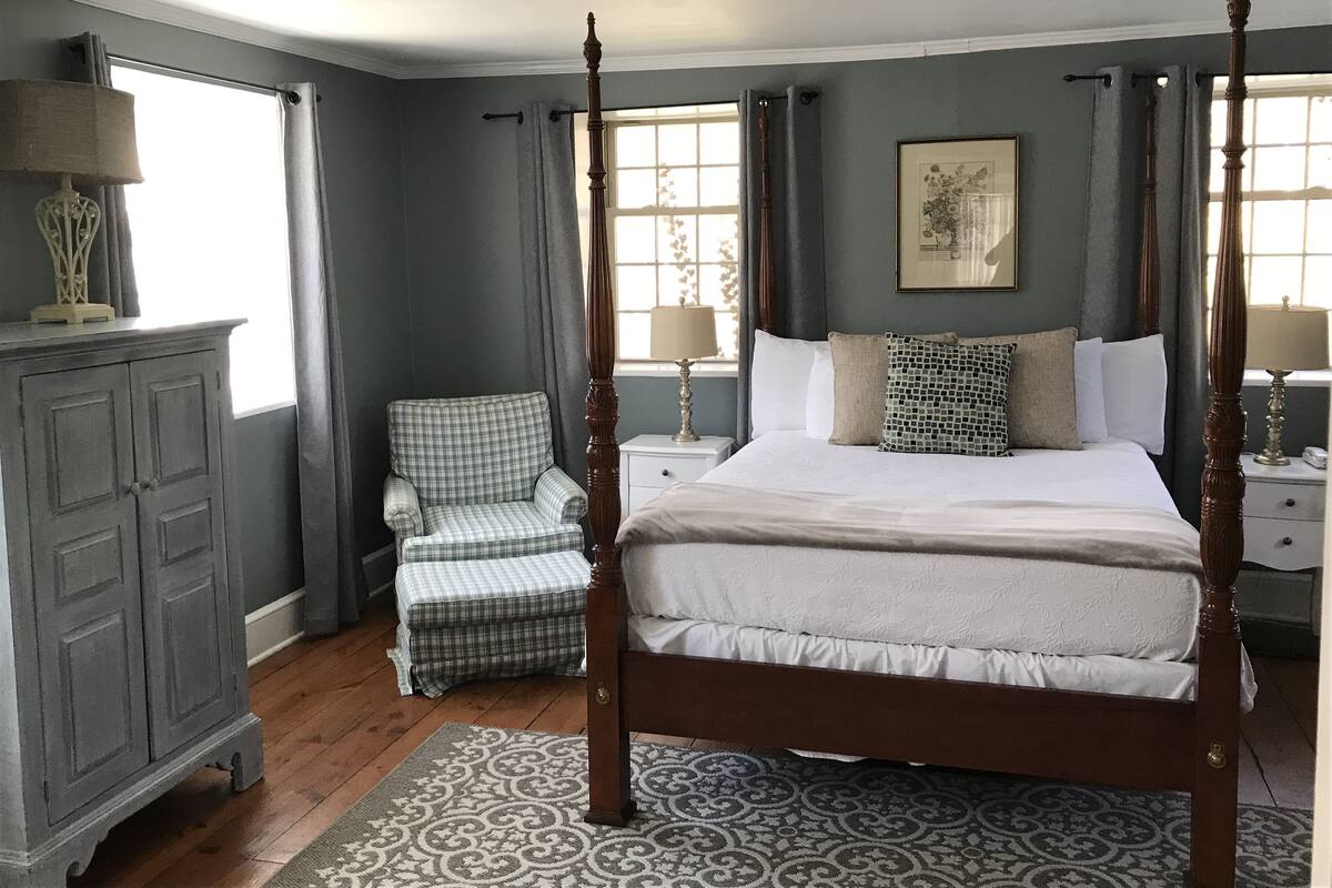 Una cama con dosel con una colcha blanca y varias almohadas, un sillón a cuadros, un aparador de madera y una alfombra estampada en un dormitorio con paredes grises y ventanas grandes.