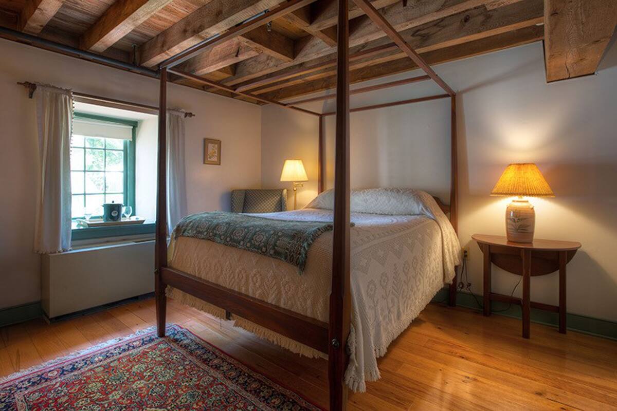 Una cama con dosel en una habitación con vigas de madera en el techo, una ventana con cortinas y dos lámparas de noche.