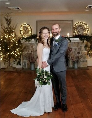 A couple in wedding attire standing in a room with wooden flooring, a fireplace, and decorative lights. - Inn at Millrace Pond (Blairstown)