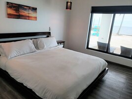 Un dormitorio con una cama grande, una mesita de noche, una lámpara de pared y vistas a la playa a través de amplios ventanales.