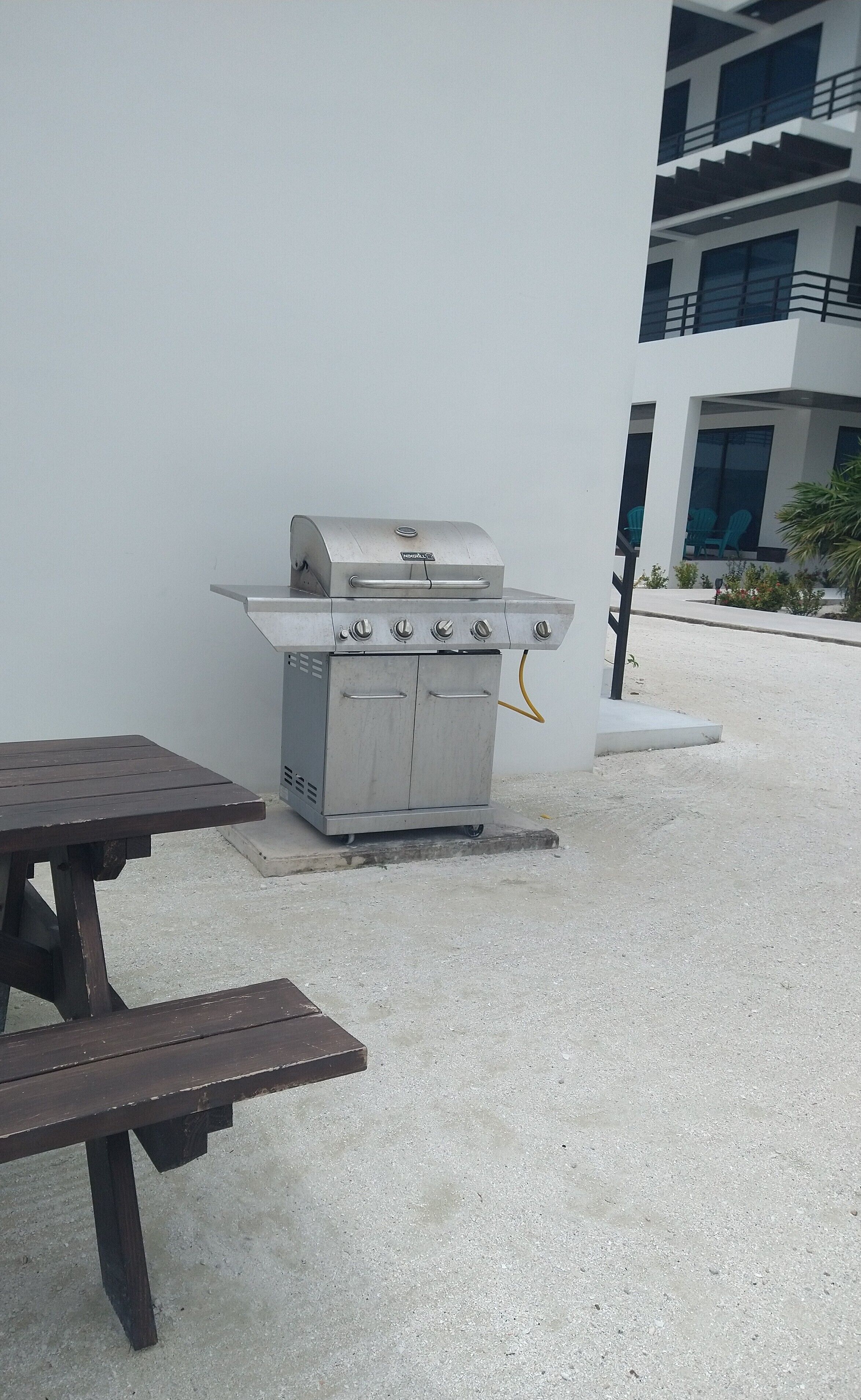 An outdoor grill with a table and benches, set against a white wall with a building in the background.