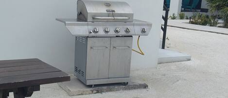 An outdoor grill with a table and benches, set against a white wall with a building in the background.