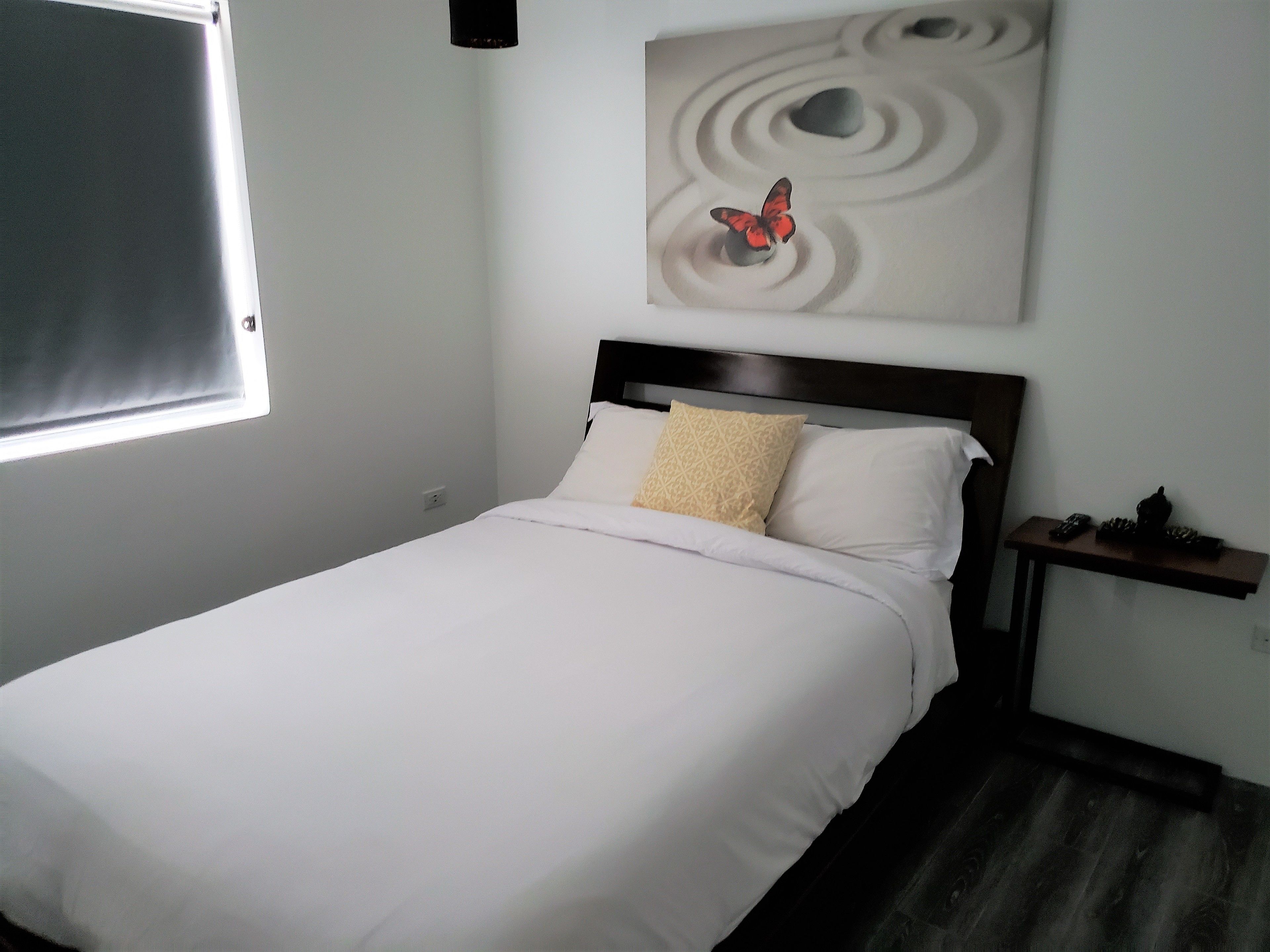 Una cama impecablemente arreglada con sábanas blancas y una única almohada estampada, una obra de arte colgada en la pared de una mariposa, y una mesita de noche con un teléfono y un objeto decorativo.