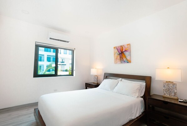 Una camera d'hotel con un letto grande, due comodini con lampade, una finestra che si affaccia su edifici e un quadro astratto dai colori vivaci appeso al muro.