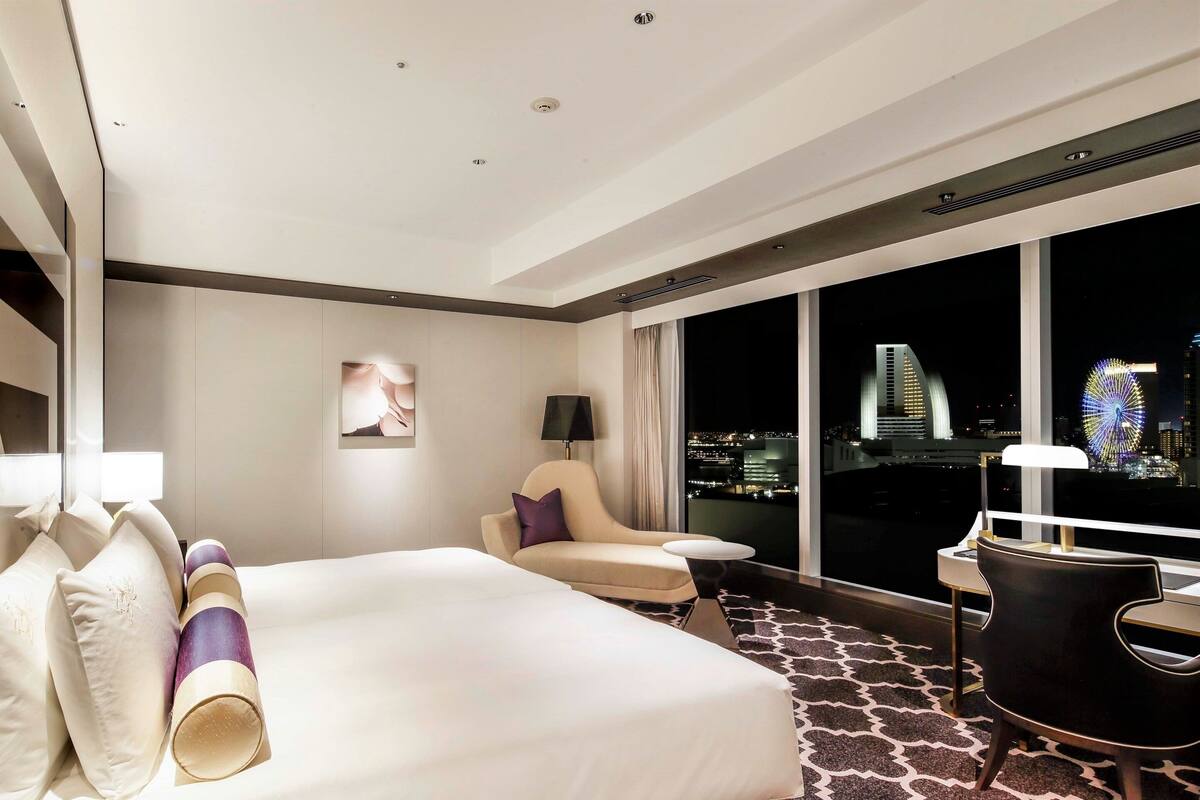 Ein Hotelzimmer mit einem großen Bett, einem Sessel, einem Schreibtisch und Blick auf die beleuchtete Stadt.