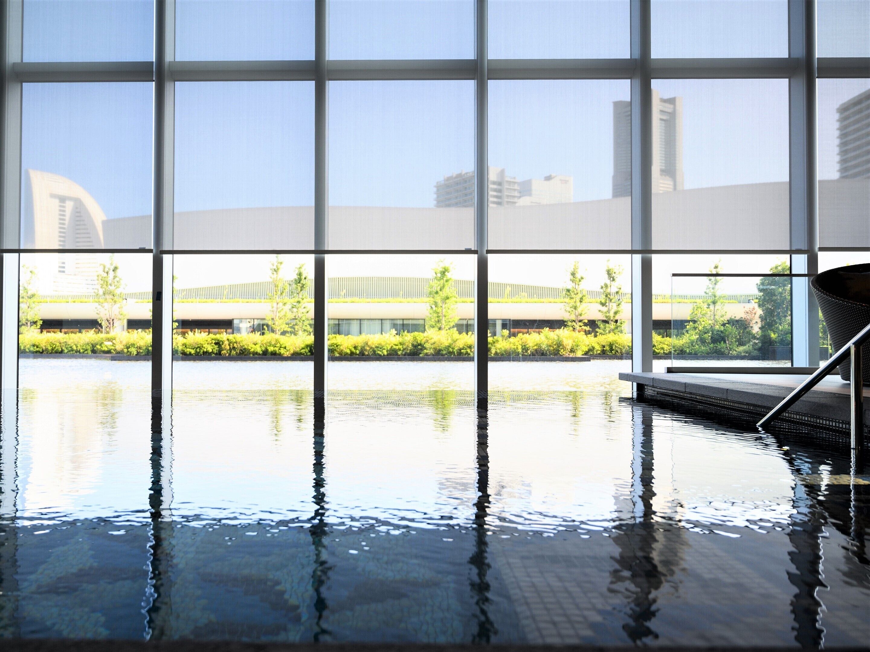 An indoor swimming pool with clear windows, a metal chair, and a view of buildings and greenery.