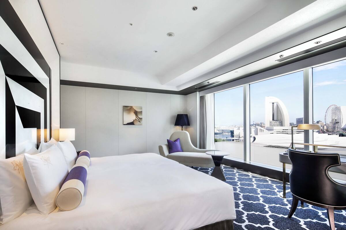 Ein modernes Hotelzimmer mit einem großen Bett, einer Sitzecke mit einem Sessel und einem kleinen Tisch sowie einem großen Fenster mit Blick auf die Stadt.
