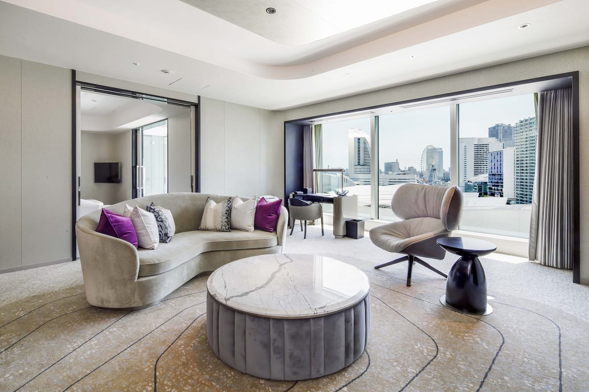 Ein modernes Wohnzimmer mit einer gebogenen Couch, einem runden Couchtisch und einem Sessel. Ein großes Fenster bietet Blick auf die Stadt.