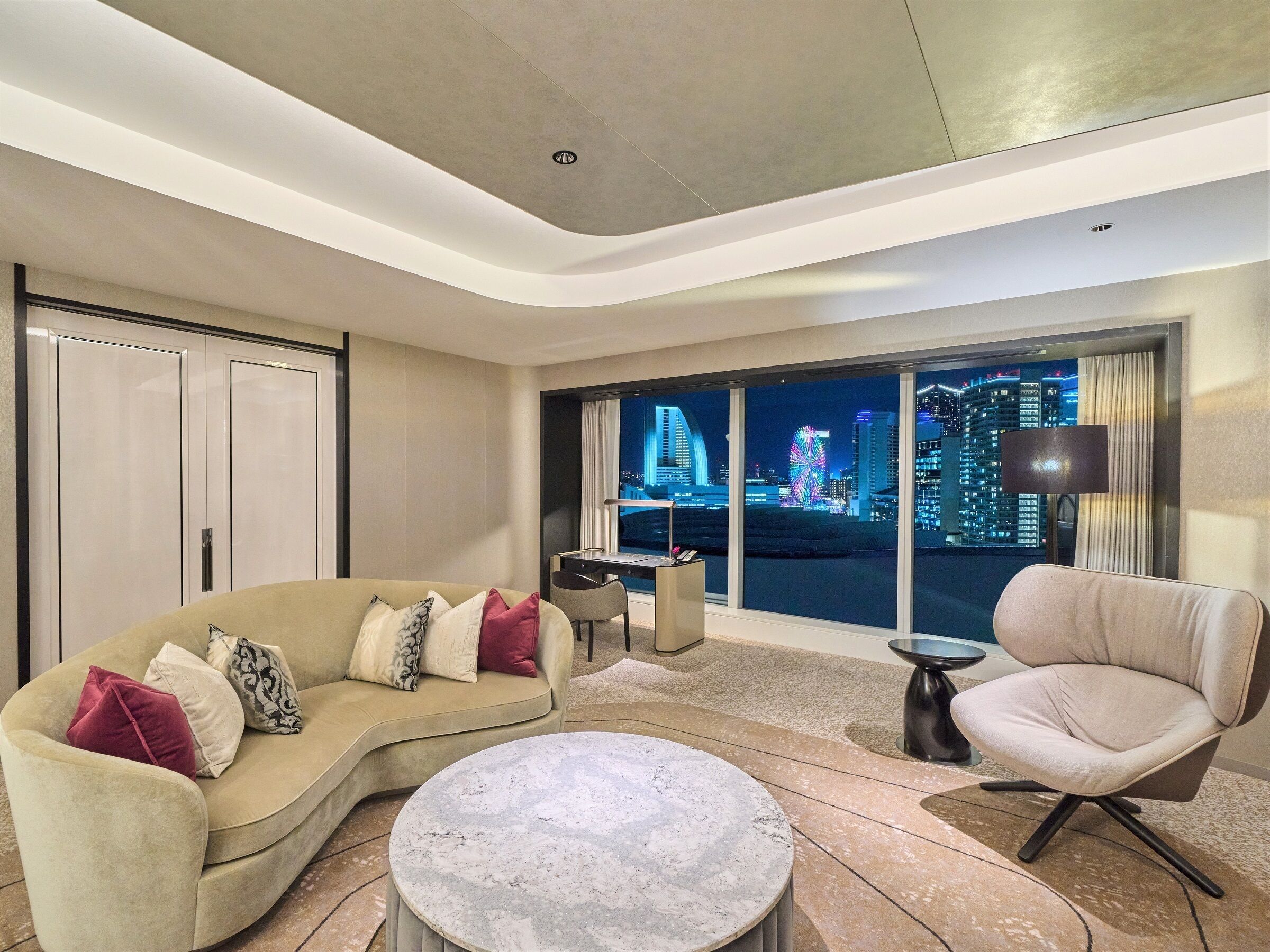 Ein modernes Hotelzimmer mit einer gebogenen Couch, einem runden Couchtisch und Blick auf eine beleuchtete Stadtlandschaft.