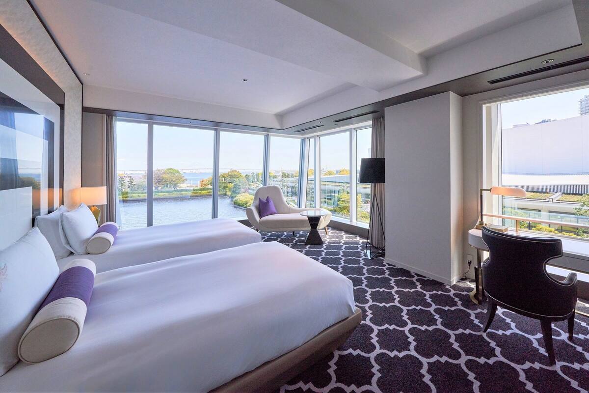 Ein Hotelzimmer mit einem großen Bett, zwei Sesseln, einem kleinen Tisch und Blick auf eine Stadtlandschaft durch große Fenster.