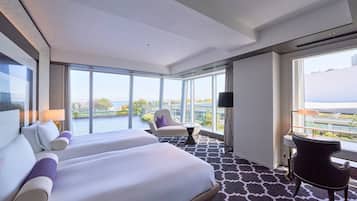 Habitación de hotel con una cama grande, dos sillones, una mesita y vistas a un paisaje urbano a través de amplios ventanales.