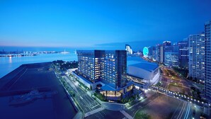 A modern high-rise hotel with a glass facade, located near a waterfront with a bridge in the background.