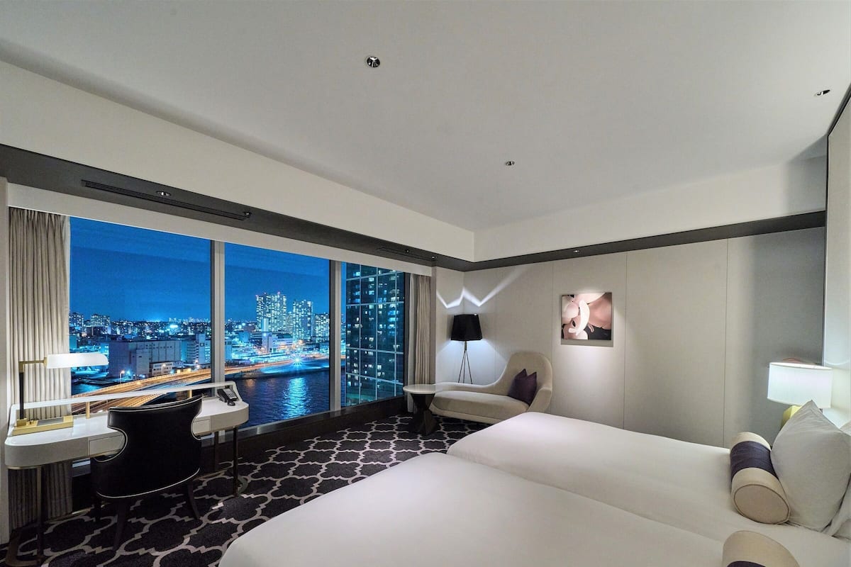 Ein modernes Hotelzimmer mit einem großen Bett, einem Schreibtisch mit Stuhl, einer Stehlampe und Blick auf eine nächtliche Skyline.