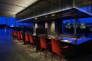 A modern bar area with a long counter, red chairs, and a view of the water and city skyline at dusk.