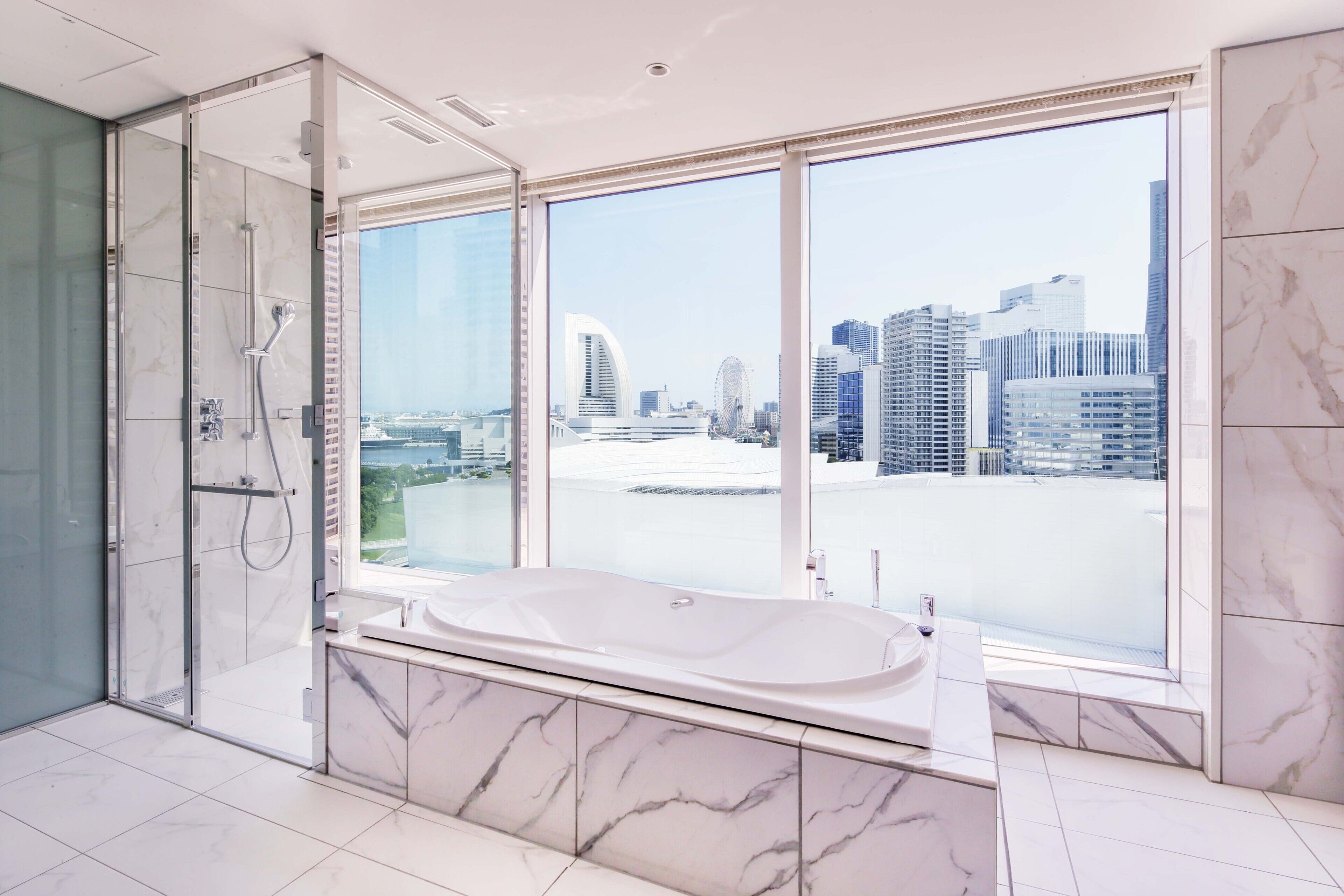 A modern bathroom with a large bathtub, a glass-enclosed shower, and a city view through large windows.
