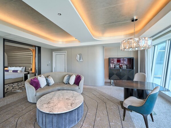 Een moderne hotelkamer met een gebogen bank, een ronde salontafel, een eettafel met stoelen, een kroonluchter en een groot raam met uitzicht.