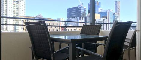 A balcony with a glass table and four chairs, overlooking a cityscape with modern buildings.